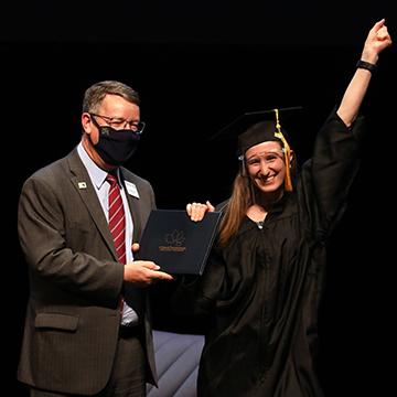 A GED graduate wearing a cap, gown and face shield receives her diploma while smiling at the camera and raising a fist in the air