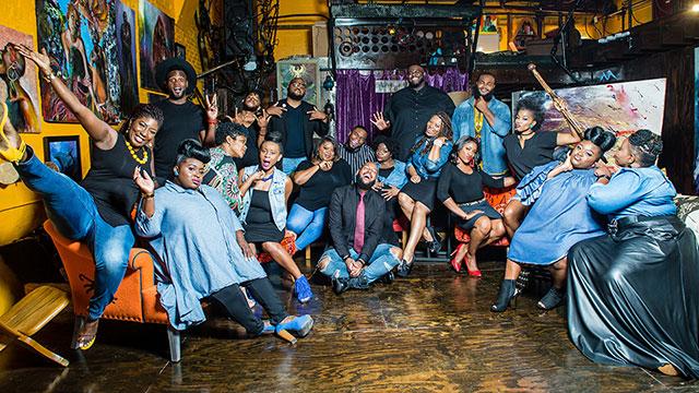 a large, celebratory group of musicians poses in an art studio with yellow walls and many art works surrounding them