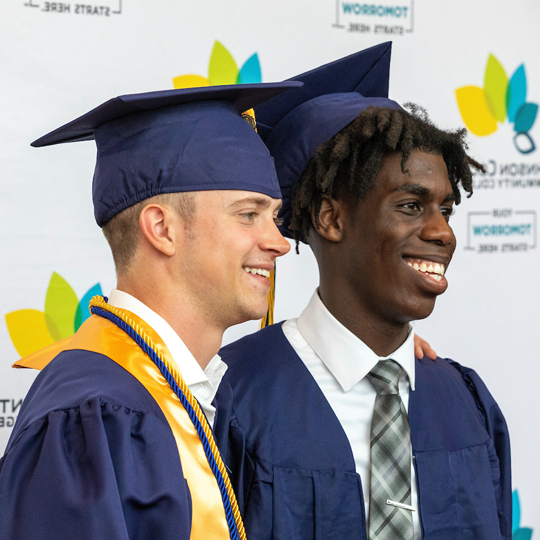 Two graduates pose for a photo in front of a backdrop that has 赌钱app可以微信提现 logos on it